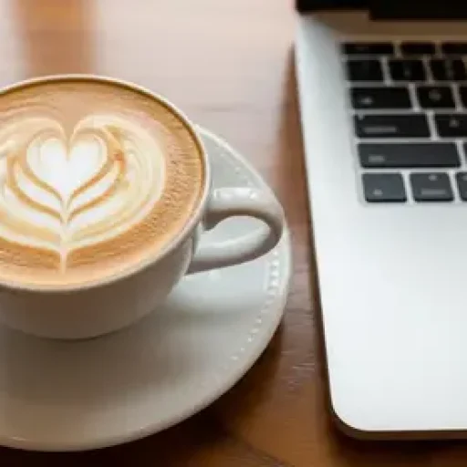 A close-up of a latte with heart art on a saucer next to a laptop on a wooden table.